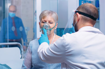 Picture of a female patient doing Respiratory Therapy.
There is a male Medical Professional holding an oxygen mask up to the female patient&apos;s mouth. He is wearing gloves, a medical mask, face shield, and a medical coat.