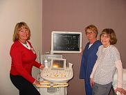 Three smiling women are standing near hospital equipment